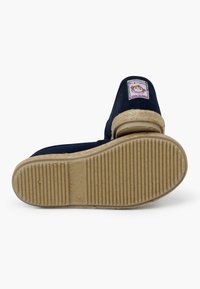 Pantofole per bambini di colore blu scuro con rivestimento in pelliccia beige, suola in gomma con striature orizzontali e un distintivo colorato a forma di scimmia sul tallone.