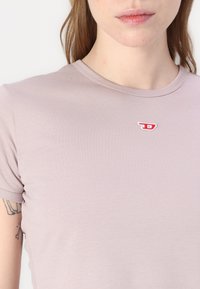 T-shirt viola chiaro realizzato in tessuto morbido e elasticizzato. Presenta un collo rotondo e un piccolo logo ricamato rosso sul petto.