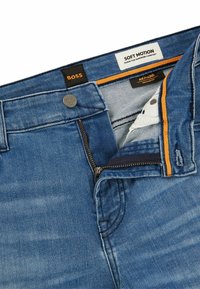 Vaqueros de mezclilla en azul medio con una textura suave, que cuentan con un cierre de cremallera, un acento naranja en el interior de la pretina y etiquetas de marca.