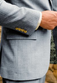 Hellgraue Sakkojacke aus strukturiertem Stoff, mit vier goldenen Knöpfen am Ärmel und einem kontrastierenden gemusterten Futter, das sichtbar ist.