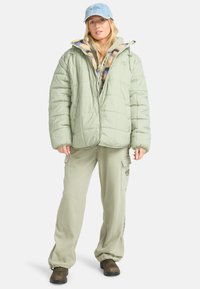 Donsachtige lichtgroene jas met een gestikt ontwerp, gedragen over een hoodie met patroon. Losvallende lichtgroene broeken en een denim pet maken de look compleet.