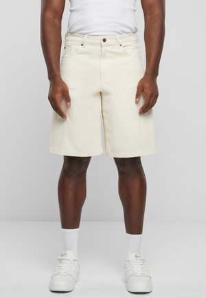 Pantaloni corti beige, lunghi fino al ginocchio, realizzati in cotone, con tasche frontali e chiusura a bottone. Indossati con una canotta bianca e sneakers.