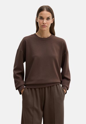 Frau mit glattem braunem Haar trägt ein lockeres braunes Sweatshirt mit langen Ärmeln und passende weite Hosen, Hände in den Taschen, neutraler Hintergrund.