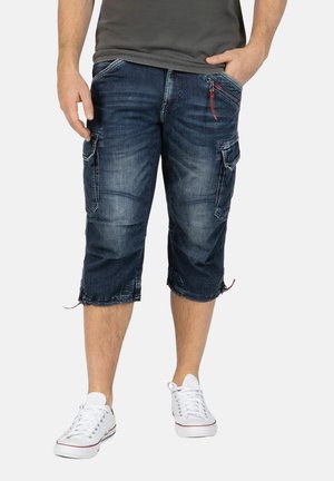 Denim cargo shorts in dark blue with knee-length cut, multiple side pockets, and a red zipper detail. Paired with white sneakers.