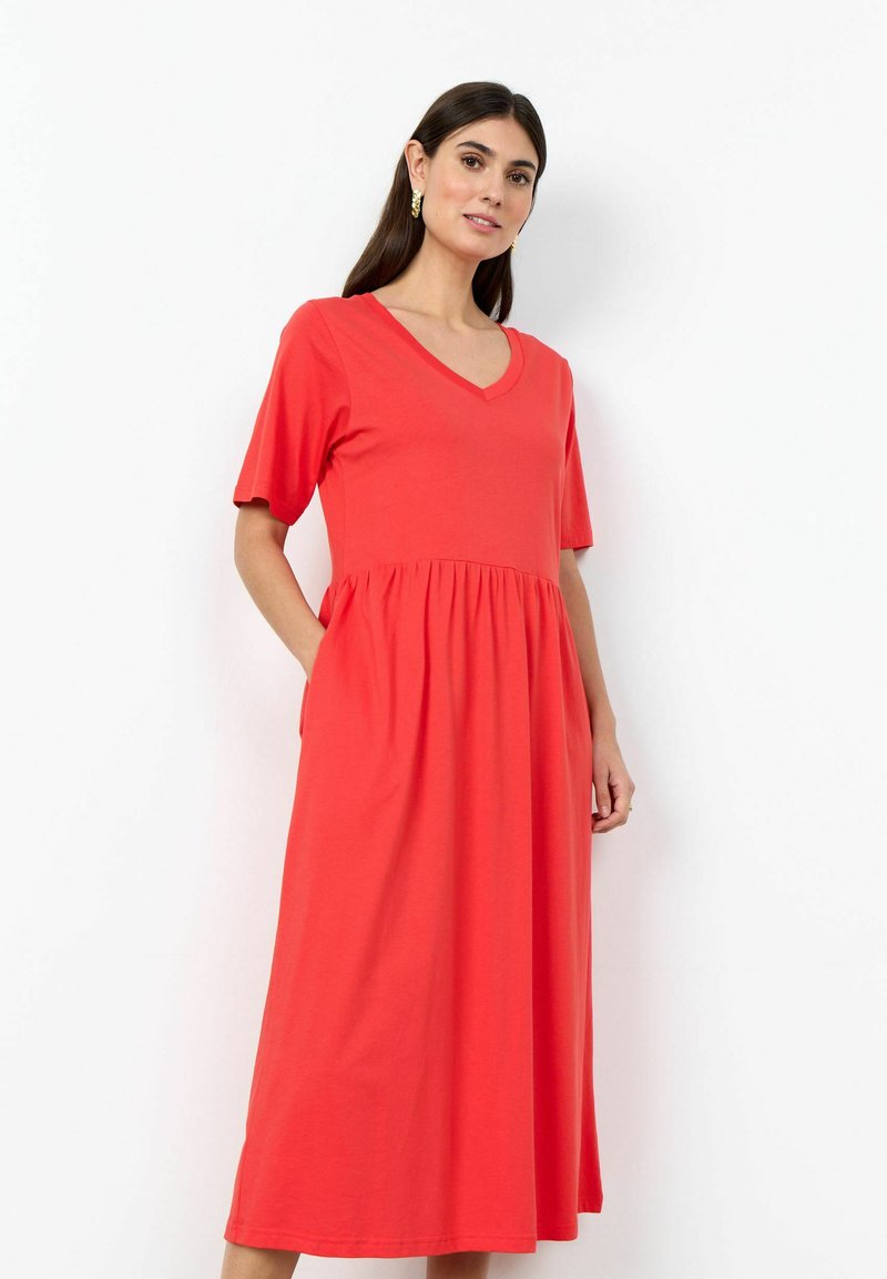 Femme portant une robe midi rouge corail à manches courtes, col en V et taille froncée, debout sur un fond blanc.