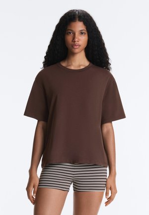 Kobieta o kręconych włosach nosi luźną brązową koszulkę z krótkim rękawem i czarno-białe krótkie spodenki w poziome paski, stoi na jednolitym tle.