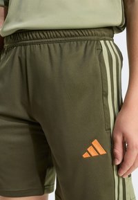 Green athletic shorts made of smooth fabric, featuring side stripes in lighter green, an orange Adidas logo, and an elastic waistband.
