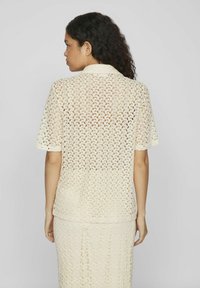 Top de color crema, de manga corta, que presenta un patrón de encaje con motivos florales. Textura suave, con un cuello tipo polo y un corte holgado.