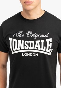 Crna majica s kratkim rukavima s odvažnim bijelim tekstom "The Original Lonsdale London" u kombinaciji rukopisnog i bloknog fonta. Materijal: pamuk.