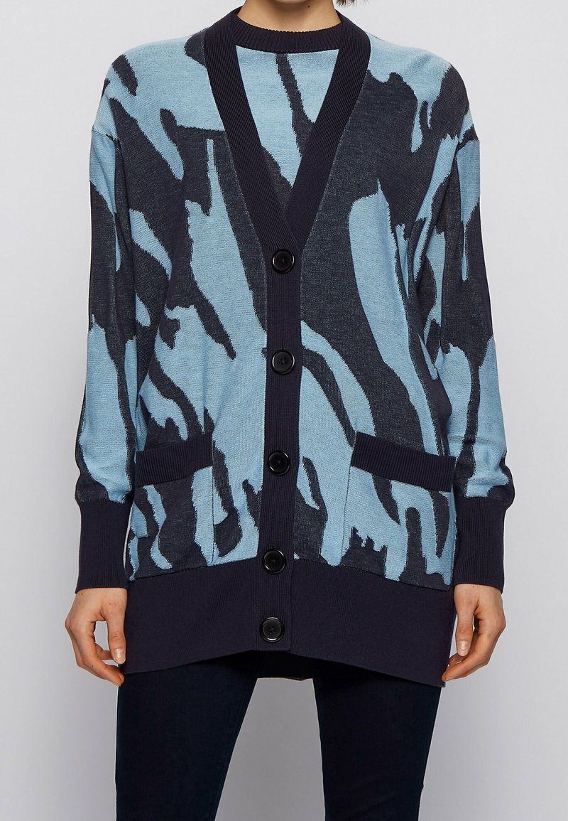 Cardigan boutonné bleu clair avec motif abstrait bleu marine foncé, col côtelé et deux poches devant ; tissu doux en maille.