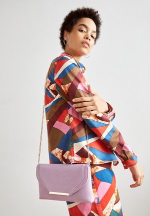 Femme portant une tenue colorée à motifs géométriques avec des boucles d’oreilles anneaux, portant un sac bandoulière violet clair avec une chaîne dorée.