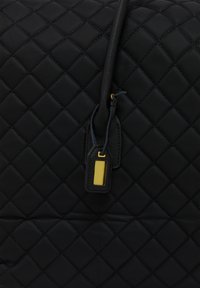 Sac en nylon noir matelassé avec une surface texturée, comprenant un tirette de zipper et un accent doré sur une petite étiquette.