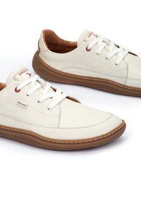 Des baskets en cuir blanc avec un bout arrondi, une semelle brune contrastante et des lacets blancs ; présentant des coutures minimales et un petit logo.