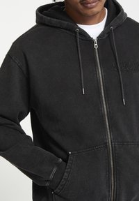 Sudadera con cremallera negra hecha de un material suave, con cremallera frontal y capucha con cordón. Incluye bolsillos laterales y un sutil bordado decorativo.