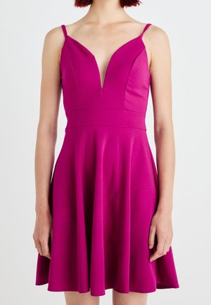 Femme portant une robe fuchsia sans manches avec un décolleté en V profond et une jupe évasée, debout devant un fond uni.
