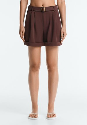 Femme portant un short marron taille haute avec ceinture et revers, associé à des sandales blanches à enfiler, debout devant un fond clair.