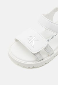 Bijele sandale s teksturiranim trakama, srebrnim "CK" logotipom i mekanom unutrašnjošću, fleksibilnom gumenom potplatom i podesivim zatvaranjem.
