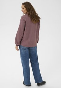Femme aux longs cheveux bruns portant un chemisier mauve à manches longues, un jean large bleu et des chaussures noires, debout devant un fond blanc.