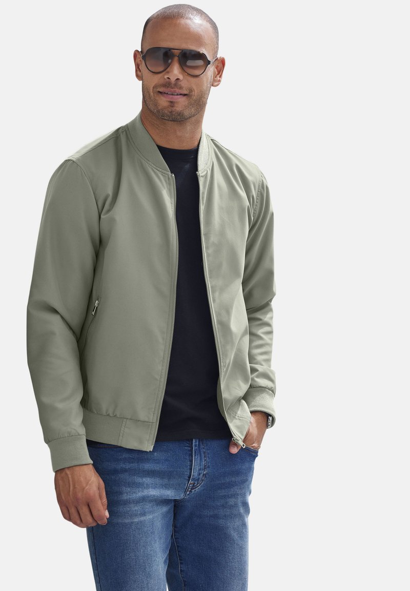 Olivgrüne Bomberjacke mit Reißverschluss, gerippten Bündchen, kombiniert mit einem schwarzen Shirt und blauen Jeans. Das Modell trägt Sonnenbrillen.