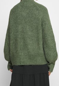 Maglione verde lavorato a maglia con un motivo testurizzato a coste. Caratterizzato da un colletto alto, spalle scese e una vestibilità comoda, in contrasto con una gonna nera.