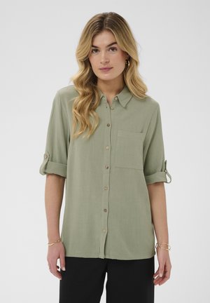 Žena s dlhými vlnitými blond vlasmi nosí svetlozelenú košeľu na gombíky s vyhrnutými rukávmi a čierne nohavice na jednoliatom svetlom pozadí.