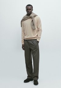 Beżowy sweter robiony na drutach nałożony na taupe'owy szalik, w połączeniu z brązowymi spodniami w paski i ciemnymi loafersami. Gładkie tekstury i luźny krój.