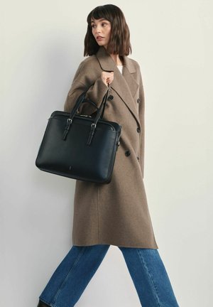 Kobieta idąca, ubrana w brązowy dwurzędowy płaszcz i niebieskie dżinsy, niesie dużą czarną skórzaną torebkę na ramieniu.