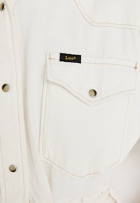 Valkoinen denim-paita, jossa on hopeiset nepparit, rintatasku mustalla tarralla ja näkyviä ompelutietoja. Teksturoitu, rento muotoilu.