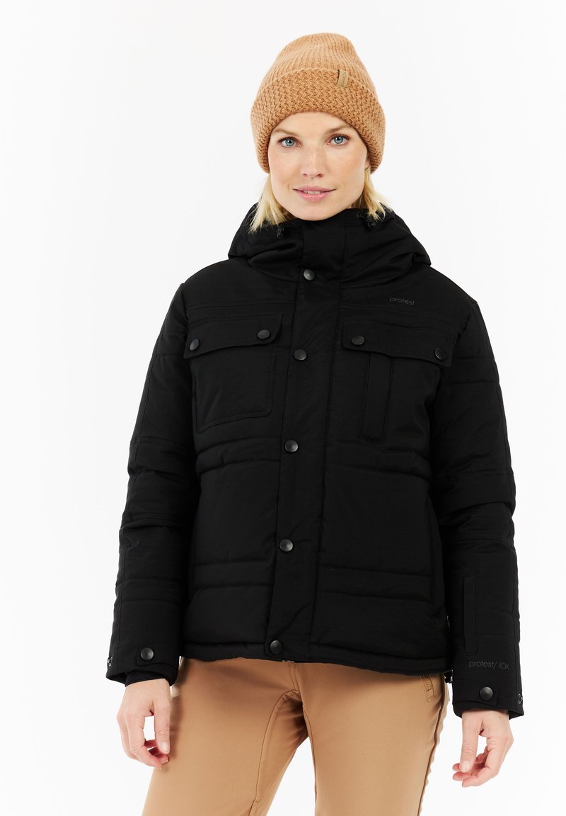 Vrouw in een zwarte winterjas met capuchon en drukknoopjes, met een bruin gebreid mutsje, staande tegen een witte achtergrond.