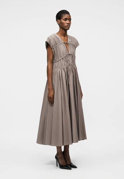 Žena stojaca v taupe plisovaných midi šatách s krátkymi rukávmi a detailmi na zaväzovanie, doplnených čiernymi špicatými vysokými podpätkami.