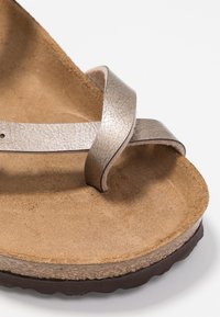 Sandală thong metalică argintie cu un breton texturat și curbat. Talpa interioară moale din plută de culoare maro și talpa exterioară din cauciuc închis, având un strop de margine ondulată.