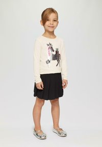 Hellecremige Langarmbluse mit einem Pailletten-Pferdemotiv, kombiniert mit einem schwarzen plissierten Rock und glänzenden silbernen Schuhen.