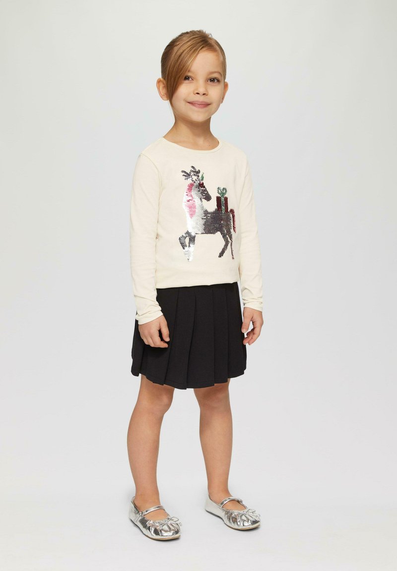 Hellecremige Langarmbluse mit einem Pailletten-Pferdemotiv, kombiniert mit einem schwarzen plissierten Rock und glänzenden silbernen Schuhen.