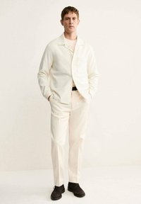 Chaqueta de camisa de corte tailored color crema con un solo bolsillo, combinada con pantalones a juego y zapatos oscuros. Textura de tela suave, diseño minimalista.