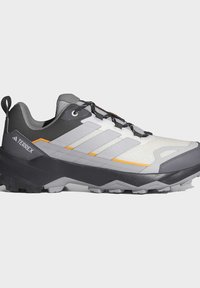 Γκρι και λευκό πεζοπορικό παπούτσι Adidas Terrex με μαύρα κορδόνια, υφή στη σόλα και πορτοκαλί γραμμές σε απλό φόντο.