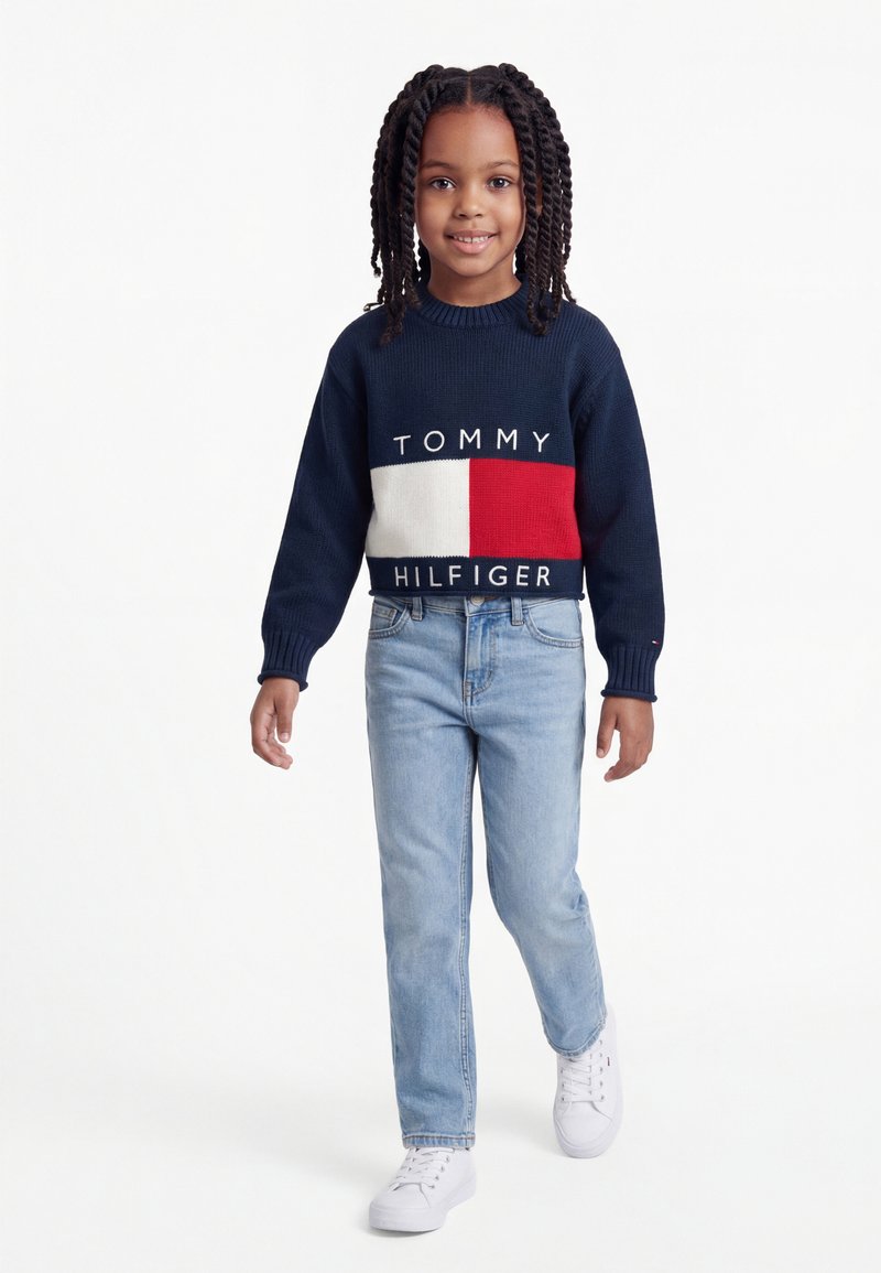 Jauna mergina su pintais plaukais, dėvinti tamsiai mėlyną Tommy Hilfiger megztinį, šviesiai mėlynas džinsus ir baltus sportbačius, žengia pirmyn ant balto fono.