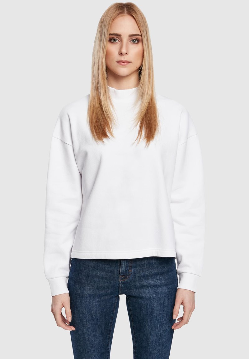 Mujer joven con cabello largo rubio que lleva un sudadera blanca simple y vaqueros azules, de pie contra un fondo gris claro liso.