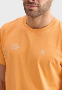 Camiseta de algodón naranja con cuello redondo, que presenta un texto blanco que dice "AUTÉNTICA MARCA DEPORTIVA DESDE 1939" y un logotipo de flor.
