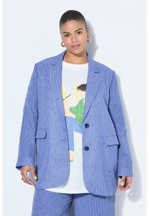 Personne aux cheveux courts portant un blazer pailleté bleu à rayures fines et un pantalon assorti, un t-shirt graphique blanc, un fard à paupières jaune et des boucles d'oreilles créoles.