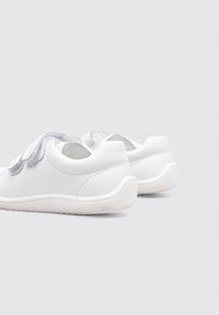 Baskets blanches en cuir avec une texture lisse, dotées d'une sangle velcro et d'un bout arrondi. La semelle est blanche avec un léger motif de crampons.