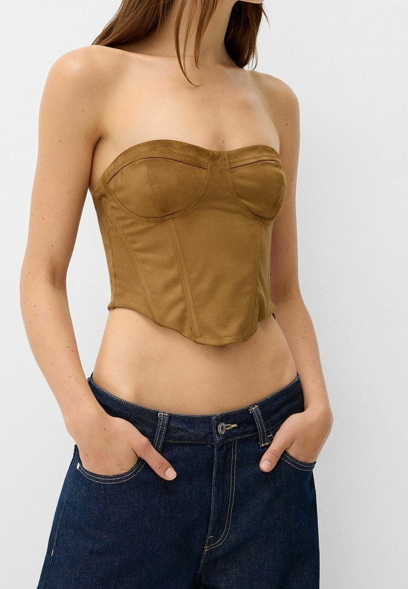 Donna che indossa un top corsetto marrone senza spalline e jeans in denim blu scuro, con le mani nelle tasche, su uno sfondo bianco semplice.
