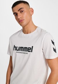Vit Hummel T-shirt med en stor svart logo och text på framsidan, korta ärmar med svarta chevron-accenter på vänster axel.