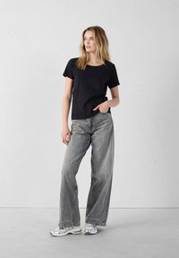 T-shirt nera in cotone con scollatura a girocollo abbinata a jeans grigi sbiaditi e dalle linee morbide. Il modello indossa sneakers argentate.