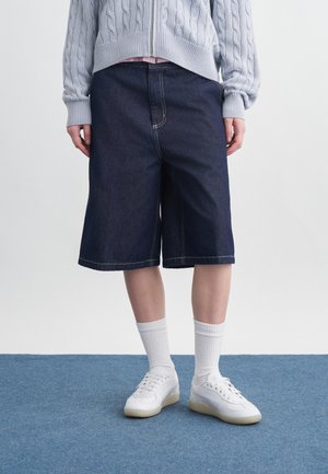 Isik, kes kannab tumel siniseid laia säärega teksapükse, valgeid pöidlaid sokke, valgeid tossusid ja helehallit palmikutega kampsunit.