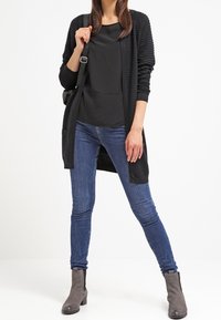 Schwarze gerippte lange Strickjacke, schwarzes Top, blaue enge Jeans und graue Stiefeletten. Einfaches Design, weicher Stoff, mit einer lässigen Silhouette.