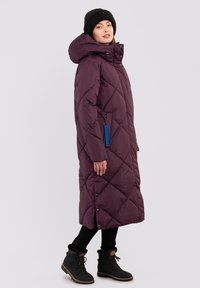 Lange, geïsoleerde pufferjas in donkerpaars met een quiltpatroon, een comfortabele capuchon en een blauwe accenttag. Gekleed met zwarte leggings en laarzen.