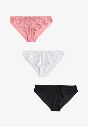 Frau mit langen dunklen Haaren, die eine weiße, geknöpfte Strickjacke, einen weißen Spitzen-BH und passende Spitzenunterwäsche trägt, steht vor einem einfarbigen Hintergrund.