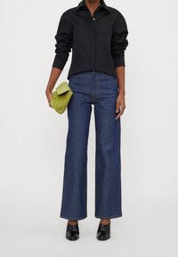 Camicia nera con bottoni, jeans in denim blu a vita alta e gamba larga, mule nere con tacco, mentre tiene una pochette verde con texture.