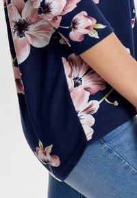 Marinblommig topp med trekvartslånga ärmar; har stora rosa blommor och gröna blad; mjuk textur; matchad med ljusblå jeans.