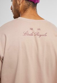 Iškilusi rožinė medvilninė marškinė su laisvu siluetu. Ant nugaros yra maroon spalvos užrašas "Étoile Royale" ir mažesni teksto akcentai.